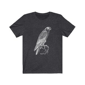 May include: A dark heather grey t-shirt featuring a white outline drawing of a falcon perched on a rock.