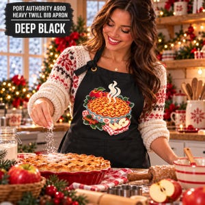 May include: A deep black Port Authority A900 heavy twill bib apron featuring a graphic print of a steaming lattice-top apple pie with cinnamon sticks and red berries. The apron has a neck strap and a front pocket.