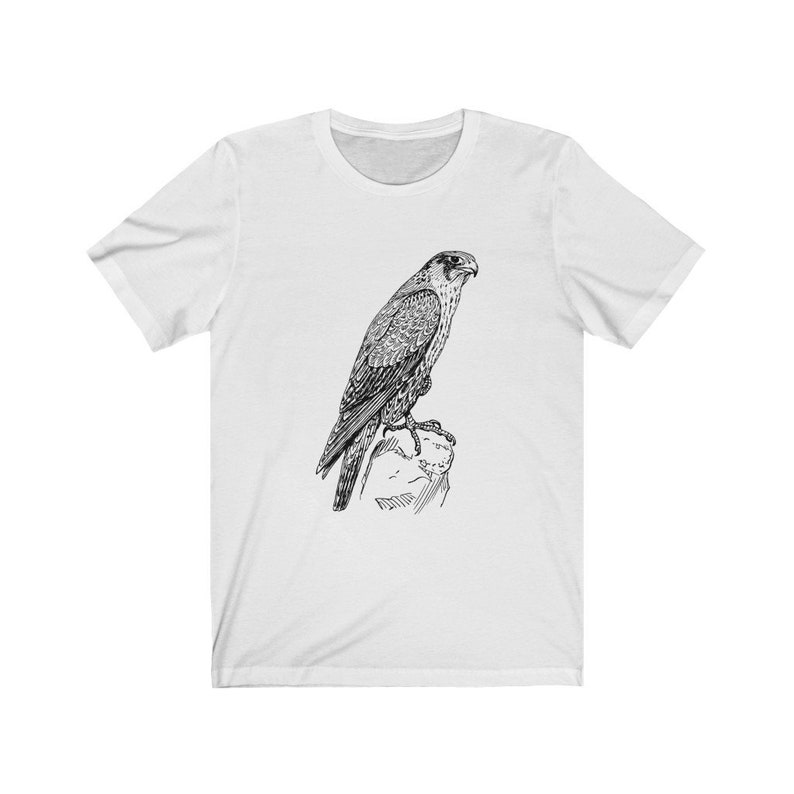 May include: A white t-shirt featuring a black and white line drawing of a falcon perched on a rock.