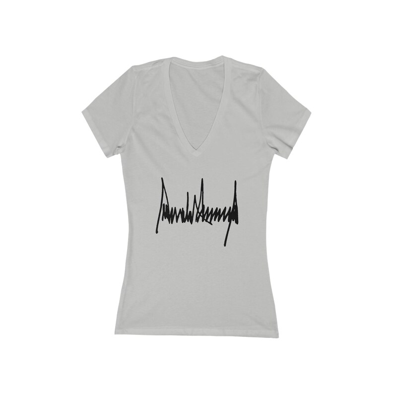 May include: White V-neck t-shirt with a black signature that reads "Donald J. Trump" printed on the front.