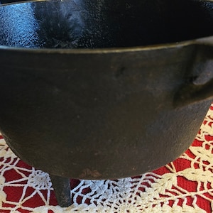 May include: A black cast iron pot with three legs and a handle. The pot is sitting on a white crocheted doily.