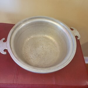 May include: A silver aluminum pot with two handles. The pot is empty and has a worn surface.