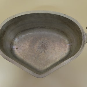 May include: A silver-colored, oval-shaped metal bowl with two decorative handles. The bowl has a smooth interior and a textured exterior on the handles. The handles are curved and have a dotted pattern. The bowl is empty.