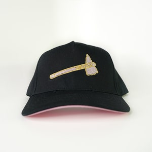 May include: Black baseball cap with a pink brim. The cap has an embroidered tomahawk design in gold and lavender. A classic style hat, suitable for casual wear.