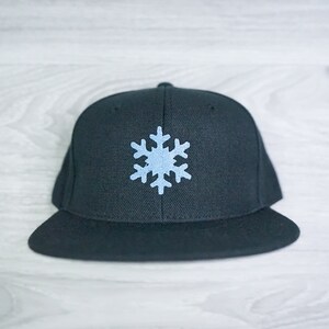 Puede incluir: Una gorra de béisbol negra con un copo de nieve azul claro bordado en la parte delantera.