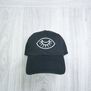 Puede incluir: Una gorra de béisbol negra con un diseño de ojo bordado en blanco.