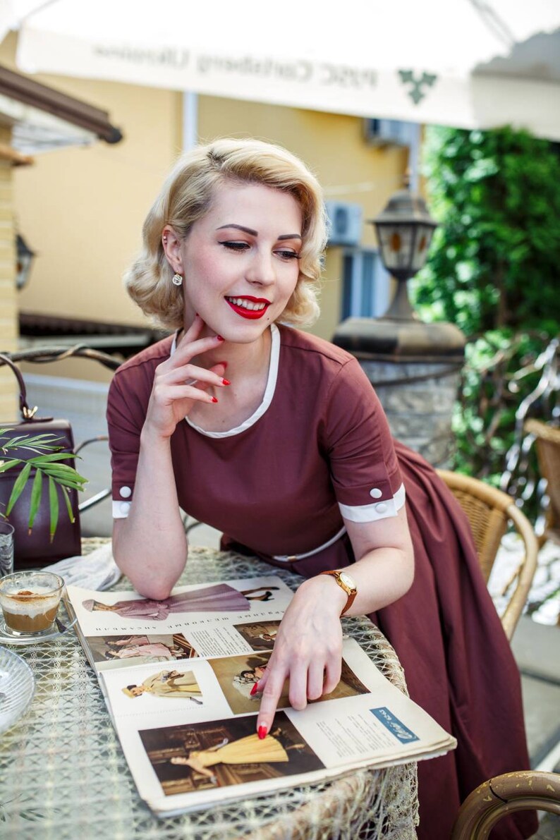 Puede incluir: Una mujer con un vestido granate con lunares blancos y ribetes blancos est&aacute; sentada en una mesa mirando una revista. Est&aacute; se&ntilde;alando una imagen de una mujer con un vestido amarillo. La revista est&aacute; abierta en una p&aacute;gina con el texto "Seasons in Vogue".