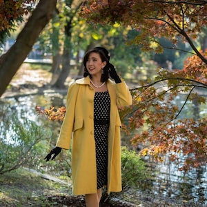 May include: A woman wearing a yellow coat over a black and white polka dot dress. She is standing in a park with fall foliage in the background.