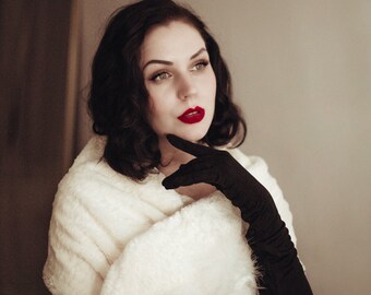 May include: A woman with dark hair and red lipstick wearing a cream-coloured faux fur shawl and long black gloves. The shawl has a decorative beaded trim. She is looking off to the side.