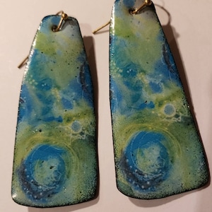 May include: A pair of long, rectangular earrings with a blue, green, and white abstract design. The earrings have a gold-colored wire hook.