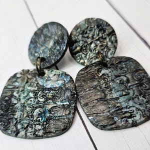 May include: A pair of handcrafted earrings with a unique, textured design. The earrings feature a dark, mottled color scheme with shades of gray, blue, and hints of gold. The design includes a circular top and a square bottom, with an ornate, swirling pattern.