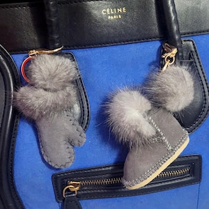 May include: A blue, black, and gray handbag with two gray fur keychains. One keychain is a mitten, and the other is a boot. The handbag has a gold zipper and the brand name "CELINE PARIS" printed on the top.