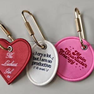 May include: Three embroidered keychains with gold-colored clasps. One is heart-shaped and red, with the text "The Louchiest Girl." The second is oval-shaped and white, with the text "I cry a lot but I am so productive." The third is pink and round, with "Main Character Energy."