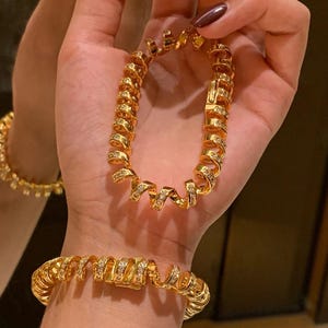 May include: Gold-toned spiral bracelets with embedded clear crystals. One bracelet is clasped on a wrist, while another is held up. The bracelets have a textured, coiled design, reflecting light. The jewelry is displayed against a neutral background.