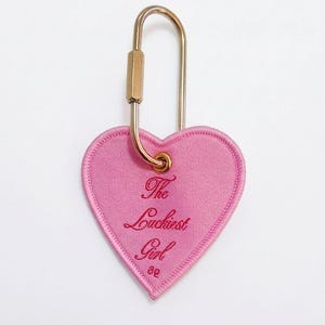 May include: A pink heart-shaped keychain with the embroidered text "The Luckiest Girl" in a cursive font. The keychain has a gold-colored metal clasp.