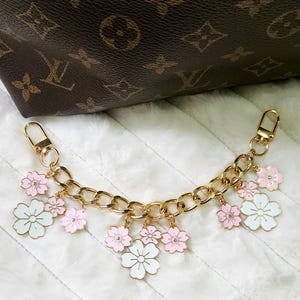 May include: Gold-tone chain bag charm with pink and white flower charms. The chain has a lobster clasp on each end for easy attachment. The flowers have a white or pink enamel finish.