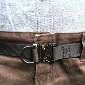 May include: Black tactical belt with a D-ring and a quick-release buckle. The belt is made of durable black nylon webbing and is fastened on brown pants. A light blue shirt is visible in the background.