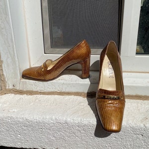 Può includere: Un paio di décolleté marroni in pelle strutturata, con punta quadrata e tacco a blocco. Le scarpe hanno un dettaglio color oro sulla tomaia e il marchio "SOFIA" è visibile all'interno di una scarpa. I tacchi sono alti circa 7.6 cm.