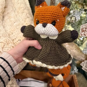 Może przedstawiać: Szydełkowa lalka lisa z pomarańczowo-brązowym futrem, ubrana w ciemnozieloną sukienkę z białym koronkowym wykończeniem. Mniejsza lalka lisa w ogrodniczkach jest widoczna w drewnianej skrzyni. Lalki mają białe pyszczki i czarne oczy.