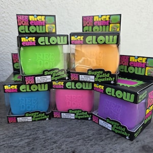 Puede incluir: Una colección de cinco juguetes sensoriales Nee Doh Nice Cube Glow en cajas individuales. Los cubos son verdes, naranjas, azules, rosas y morados. Cada caja presenta el texto Nee Doh Nice Cube Glow y Super Solid Squish en el empaque.