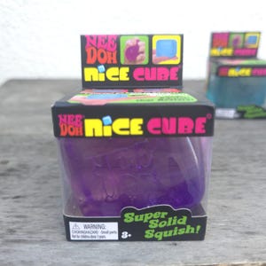 May include: A purple Nee Doh Nice Cube sensory toy inside a black cardboard box. The packaging features the text Nee Doh Nice Cube and Super Solid Squish, with a warning label about small parts and a choking hazard for children under 3 years.