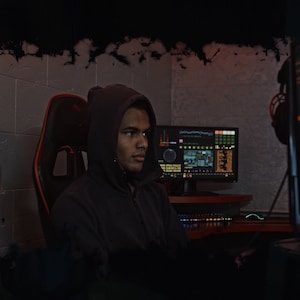 May include: A person wearing a black hooded sweatshirt sits in a gaming chair in front of a computer monitor. The monitor displays a digital dashboard with various data points. The person is looking at the monitor.