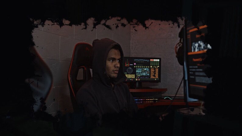 May include: A person wearing a black hooded sweatshirt sits in a gaming chair in front of a computer monitor. The monitor displays a digital dashboard with various data points. The person is looking at the monitor.