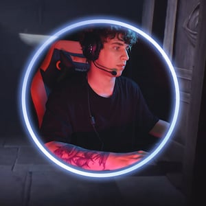 Könnte beinhalten: Eine Person mit einem schwarzen T-Shirt und Kopfhörern sitzt auf einem Gaming-Stuhl mit einem blauen Neonring um sie herum. Der Stuhl hat ein rot-schwarzes Design.