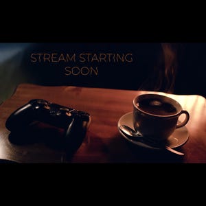 Peut inclure: Une manette de jeu noire et une tasse de café sur une table en bois. La tasse de café est posée sur une soucoupe avec une cuillère. Le texte "STREAM STARTING SOON" est affiché en orange.