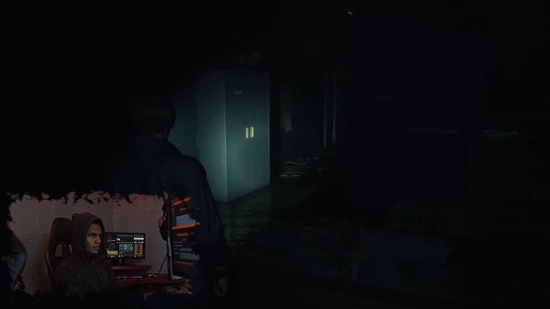 May include: A person wearing a hooded sweatshirt sits in front of a computer screen with a dark background. The screen displays computer code. The person is looking at the screen.