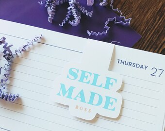 Op de afbeelding: Een blauwe en witte sticker met de tekst "SELF MADE BOSS" op een gelinieerd notitieboekblad. Het notitieboek heeft de dagen van de week en data erop geschreven. De sticker bevindt zich op de pagina voor donderdag 27.