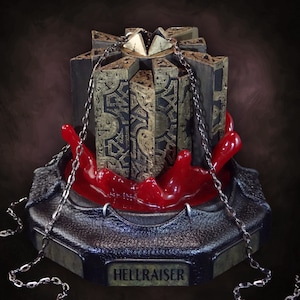 May include: A detailed sculpture of the Hellraiser puzzle box, featuring black panels with gold filigree, a gold crown, and a red, hand-shaped base. Silver chains drape around the box, and the base reads "HELLRAISER."
