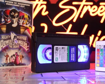 Może przedstawiać: Czarna kaseta VHS z etykietą "Mighty Morphin Power Rangers The Movie" podświetlona niebieskimi i fioletowymi światłami. Pilot z opcjami kolorów znajduje się po prawej stronie. Po lewej stronie znajduje się pudełko po filmie VHS.