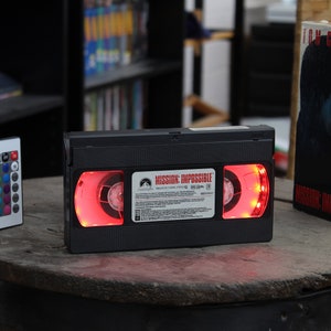 May include: A black VHS tape with the title "Mission: Impossible" illuminated with red lights. A remote control with color options sits to the left. The movie box is visible to the right.
