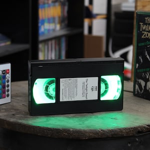 May include: A black VHS tape with illuminated green reels, labelled "The Twilight Zone." A colour-changing remote control is next to it. A VHS box of "The Twilight Zone" is also present, suggesting a retro theme.