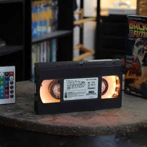 Puede incluir: Una cinta VHS negra con la etiqueta "Regreso al Futuro" está iluminada, con una caja de película a juego al lado. Un pequeño mando a distancia con botones multicolores está a la izquierda. Los objetos están sobre una superficie de madera.