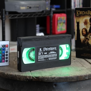 May include: A black VHS tape of "The Lord of the Rings" with green glowing reels, next to a remote control with colorful buttons and a movie box. The items are on a wooden barrel.