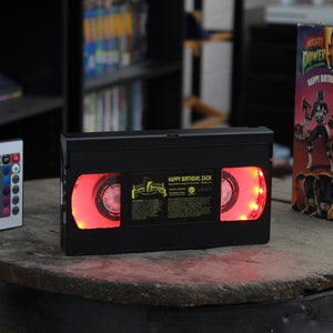 以下が含まれることがあります： 「HAPPY BIRTHDAY, ZACK」の文字が赤色に光る黒いVHSテープ。左にはカラーオプション付きのリモコンがあります。「Mighty Morphin Power Rangers」のVHSボックスが右側にあります。