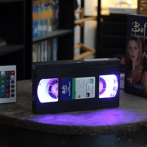 以下が含まれることがあります： 紫色の光を放つ黒いVHSテープ、カラーオプション付きのリモコン、そして「バフィー・ザ・バンパイア・スレイヤー」のDVDボックスセットが木製の表面に展示されています。VHSテープには「Buffy」と書かれたラベルが付いています。