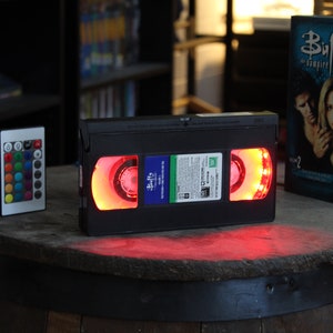 以下が含まれることがあります： 赤いLEDライトが付いた黒いVHSテープ、リモコン、そして「バフィー・ザ・バンパイア・スレイヤー」のDVDボックスセットが木製の樽の上に展示されています。VHSテープには番組タイトルが書かれた青いラベルが付いています。