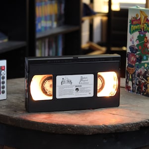 May include: A black VHS tape converted into a lamp, with glowing spools. The lamp sits on a wooden surface, accompanied by a remote control and a VHS tape box. The tape label reads "Alpha's Magical Christmas".