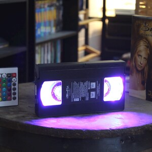 Puede incluir: Una cinta VHS negra con luz púrpura que emana de las bobinas. Un mando a distancia con botones de colores está a la izquierda. Una caja de cinta VHS con el título "Buffy Cazavampiros" está a la derecha.