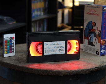 Puede incluir: Una cinta VHS negra con el título "Billy Madison" iluminada con luces rojas y amarillas. Un mando a distancia con múltiples opciones de color está a la izquierda. Una caja de cinta VHS, también etiquetada "Billy Madison", está a la derecha.