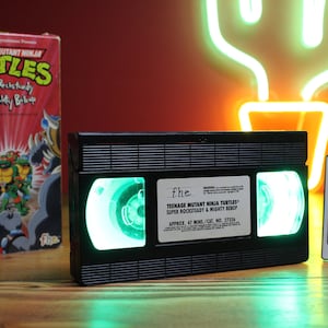 May include: A black VHS tape with green glowing reels, labeled "Teenage Mutant Ninja Turtles." A remote control with color options sits beside it. A VHS box with the same title is on the left.