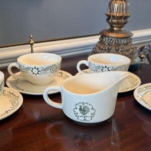 May include: A set of three cream-colored ceramic teacups with matching saucers and a creamer pitcher. Each piece features a dark green printed pattern of flowers, leaves, and geometric shapes. The creamer pitcher displays a central rooster graphic.