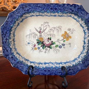 Puede incluir: Una fuente de cerámica antigua con un diseño floral azul y blanco. La fuente presenta una imagen central de un pájaro, flores y follaje. El borde tiene un patrón azul detallado. Se exhibe sobre un soporte de metal negro.