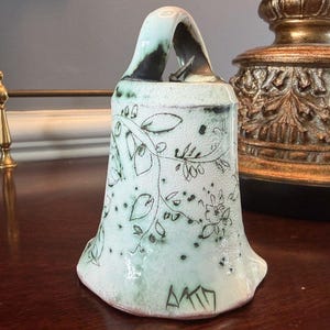 May include: A hand-formed ceramic bell with a pale mint green glaze and a dark, speckled finish. The surface features a delicate, hand-drawn black ink illustration of trailing vines, leaves, and small flowers, with a signature at the base.