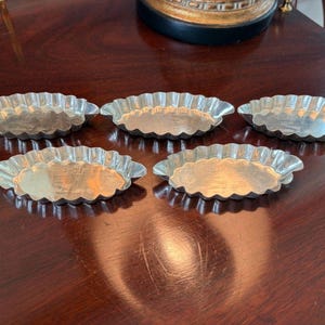 May include: A set of five oval-shaped metal tart pans with fluted edges. Each individual baking mold features a shallow basin and a scalloped rim, constructed from a shiny, silver-toned material with a smooth, reflective interior surface.