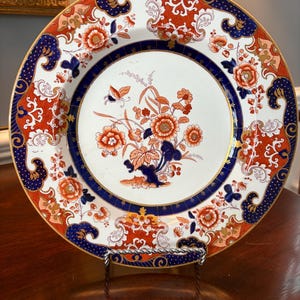 May include: A decorative plate with a white background, featuring a floral design in orange, blue, and gold. The plate has a blue border with gold accents and is displayed on a metal stand. The plate's design includes flowers and butterflies.