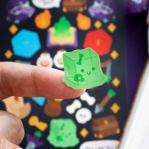 May include: A green slime cube sticker with a skull and bone inside. The sticker has a cute cartoon face with a smile and closed eyes. The sticker is on a finger.